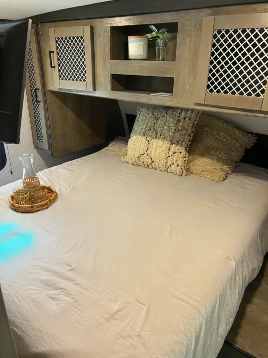 Cozy RV rental bedroom featuring clean white bedding, overhead cabinets with decor, and a convenient TV.. Heartland RVs North Trail 2021