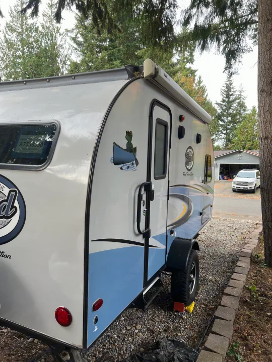 A grey and blue compact RV rental, a small travel trailer with a frog decal, is parked on gravel, ready for adventure.. Forest River R-Pod 2018