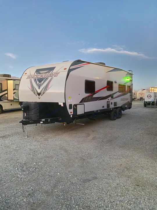 A white Shockwave toy hauler, ideal RV rental, parked on gravel. A green light glows on its rear. Other RVs are in the background.. Forest River Shockwave 2021