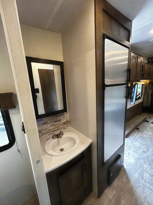 RV rental interior: Bathroom vanity with sink & mirror, adjacent to a large refrigerator & living area with TV.. Forest River Shockwave 2021