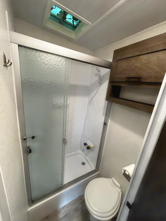 RV rental bathroom features a compact shower with glass doors, toilet, overhead fan, and wooden storage cabinet.. Forest River Shockwave 2021