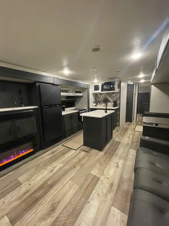 Modern RV rental interior with a well-equipped kitchen, island, and living area with an electric fireplace. Dark cabinetry.. KZ Sportsmen 2022
