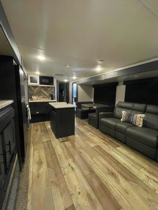 This modern rv rental interior showcases a dark kitchen island, a comfortable sofa, and dining area with light wood floors.. KZ Sportsmen 2022