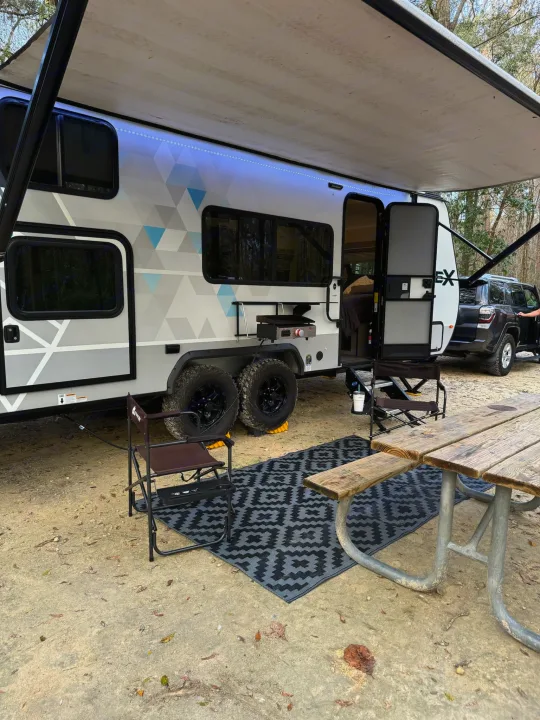 Enjoying the outdoors: an RV rental with an extended awning, chairs, rug, and picnic table on a dirt campsite.. Forest River Ibex 2021