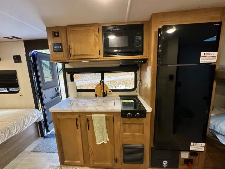 An RV rental interior showcasing a compact kitchen with a sink, stovetop, microwave, and large black refrigerator.. Cherokee Wolf pup 2025