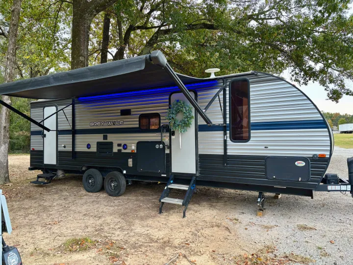 A Grey Wolf travel trailer RV rental, with a blue-lit awning and wreath, is parked at a serene campsite.. Forest River Cherokee Grey Wolf 2019