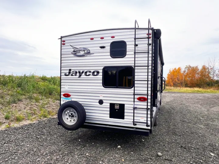 Explore with this Jayco travel trailer, perfect for an RV rental. Its rear features a ladder & spare tire; parked on gravel.. Jayco Jayflight 2021