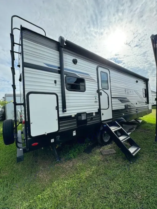 This white and gray Puma travel trailer, perfect for RV rental, features an awning, rear ladder, and steps.. Palomino Puma 2023