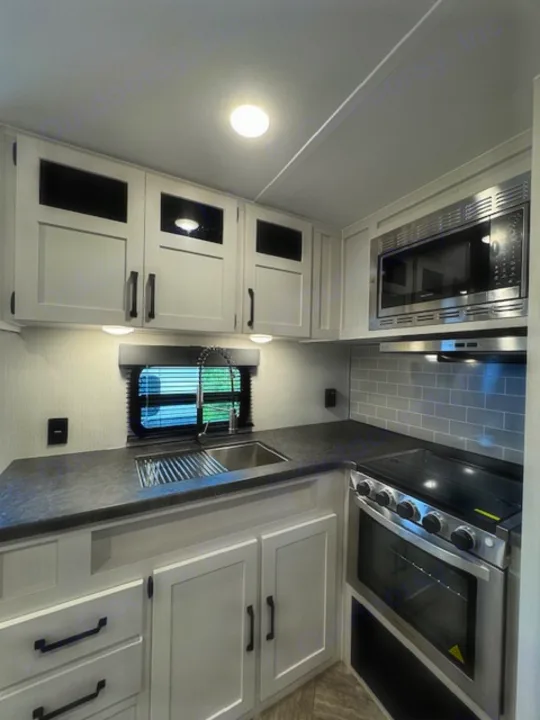 Contemporary RV rental kitchen with light grey cabinets, dark countertops, a sink, microwave, and stove/oven.. Palomino Puma 2023