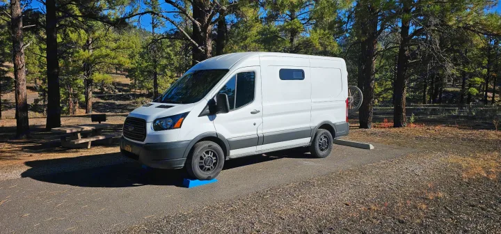 White Ford Transit camper van parked on leveling blocks at a wooded campsite with a bike rack, ideal for an rv rental journey.. FORD Ford Transit 2015