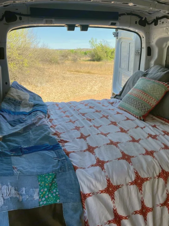 Cozy bed in an RV rental, with open back doors revealing a dry, natural landscape under a clear sky.. FORD Ford Transit 2015