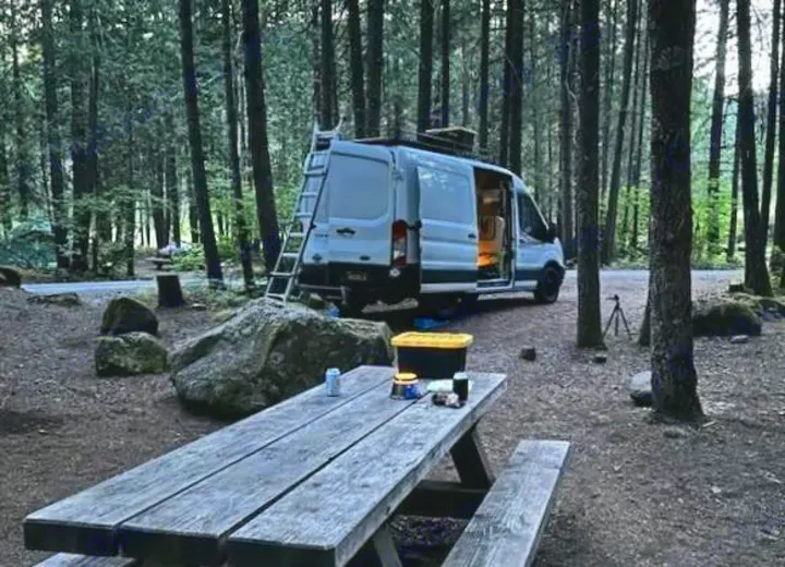 An RV rental camper van is parked with open doors at a forest campsite, alongside a picnic table with items.. FORD Ford Transit 2015