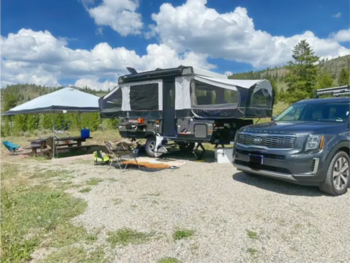 A dark SUV next to an RV rental pop-up camper, fully set up at a gravel campsite with a shaded picnic area.. Forest River Rockwood ESP 2022