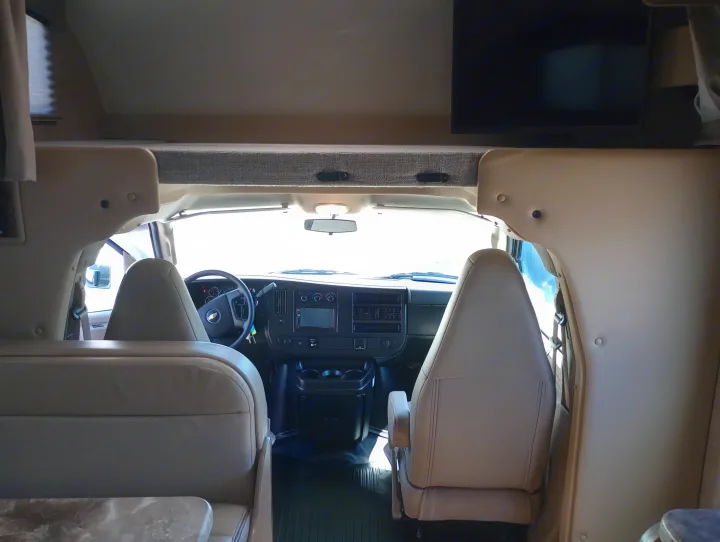 Interior view of an RV rental, showing the cockpit, front seats, and overhead bunk area with a TV.. Thor Chateau 2022