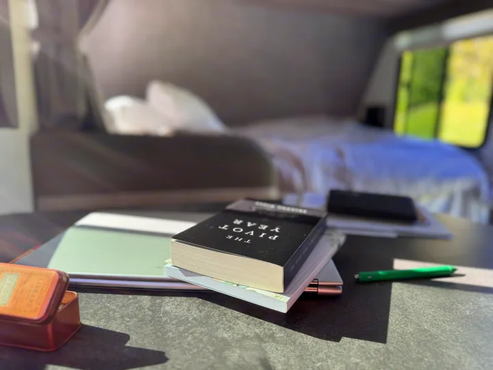 Inside an RV rental, a workspace setup with books, a laptop, and a pen on a table, with a cozy bed visible in the background.. Winnebago Minnie Winnie 2022