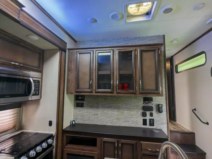 RV rental interior with a modern kitchen: microwave, stove, dark wood cabinets, and control panels are visible, with steps beyond.. K-Z Manufacturing Venom V3411TK 2020