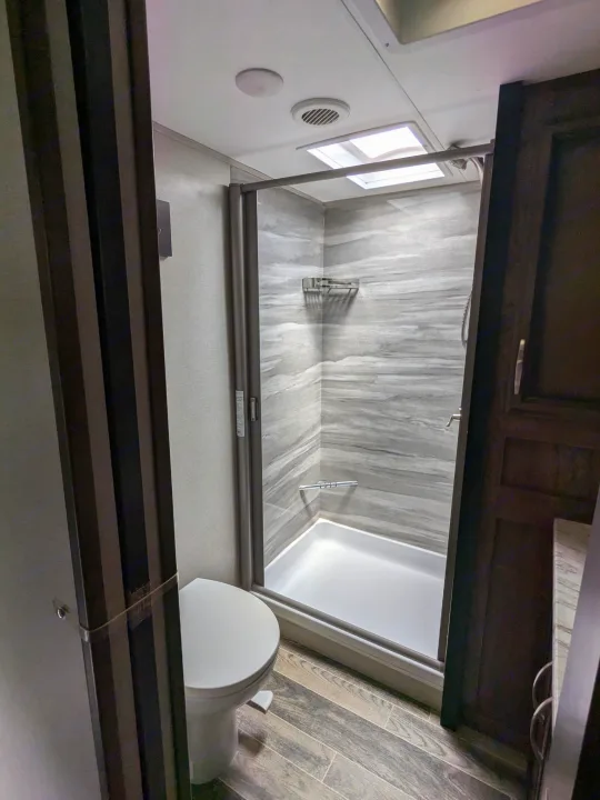 Inside an RV rental bathroom: modern shower with grey walls, skylight, white toilet, and wood-look floor visible.. K-Z Manufacturing Venom V3411TK 2020