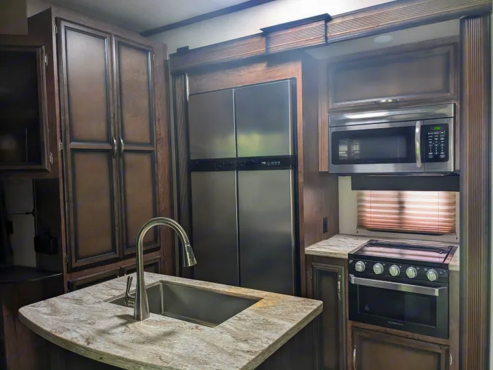Inside an RV rental: a sleek kitchen with stainless steel appliances (fridge, microwave, oven), a sink, and dark wood cabinets.. K-Z Manufacturing Venom V3411TK 2020