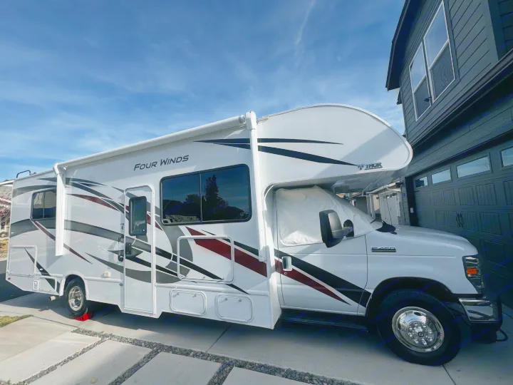 Large white RV rental parked on a driveway next to a modern house. Features sunshade, awning, and leveling blocks.. Thor Motor Coach Four Winds 2022