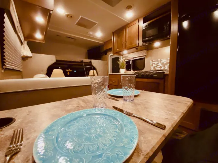 An RV rental's interior: a dining table with blue plates, a kitchen with microwave/stove, and a bed in the background.. Winnebago 25 2019