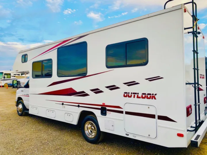 White Outlook Class C RV rental with red and maroon graphics, featuring windows and a rear ladder, parked outdoors under a blue sky.. Winnebago 25 2019