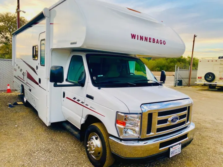 A white Winnebago RV rental, featuring a Ford front grille and side awning, is parked on gravel, ready for adventure.. Winnebago 25 2019