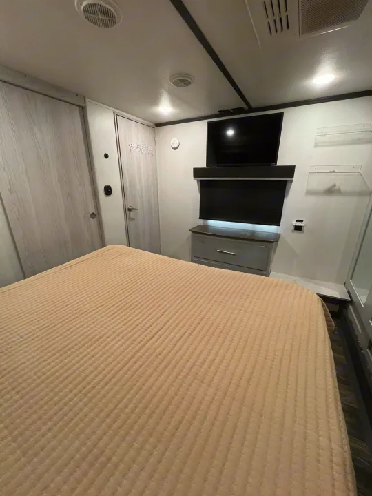 RV rental bedroom: Compact interior featuring a ribbed tan bed, gray dresser, wall-mounted TV, and light wood-grain closet doors.. Keystone RV Fuzion Toy Hauler 2021