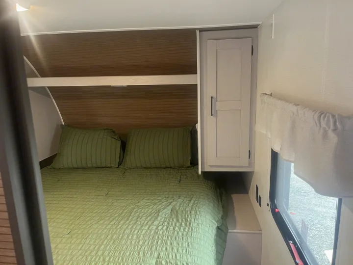 An RV rental bedroom, featuring a green-sheeted bed, light-colored overhead cabinet, and a window with a valance.. Heartland RVs Prowler Lynx 302 BHX 2025