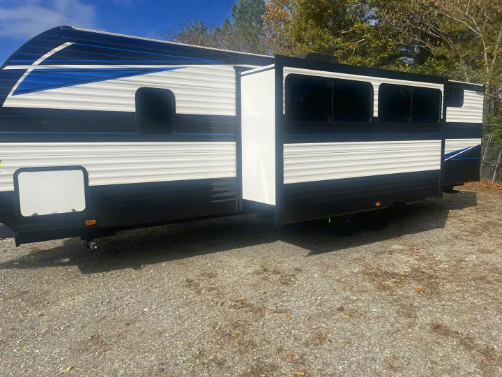 Spacious white and black RV rental with blue accents and an extended slide-out, parked on a gravel lot. Ready for your travels!. Heartland RVs Prowler Lynx 302 BHX 2025