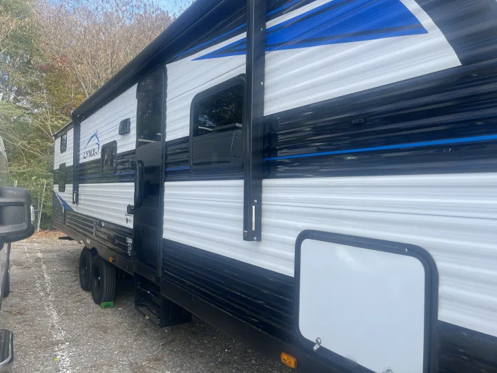 White & black Lynx travel trailer RV rental parked on gravel. Features awning, windows, and bold blue accents.. Heartland RVs Prowler Lynx 302 BHX 2025