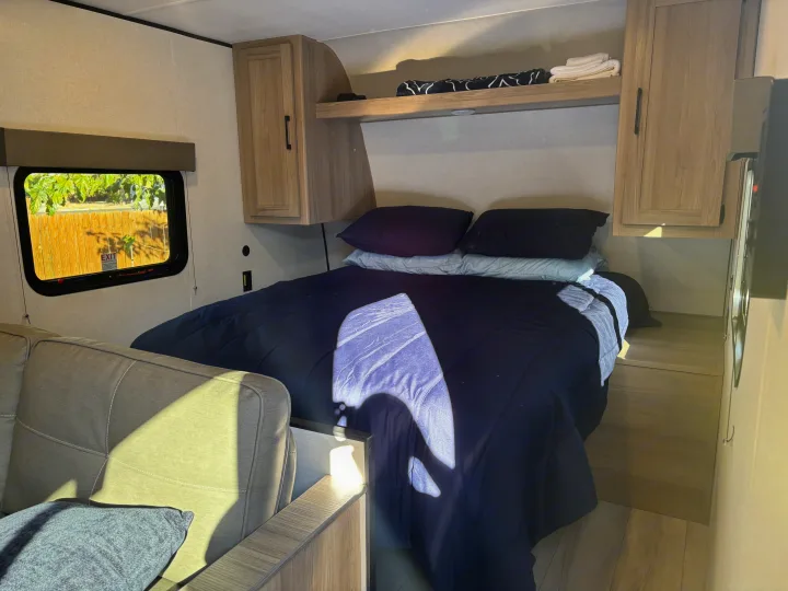 An rv rental interior shows a bed with blue bedding, overhead cabinets, and a window.. Coleman Coleman 23 B 2025