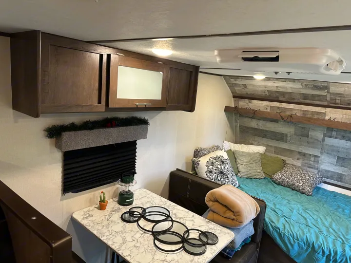 RV rental interior: cozy bedroom area with blue comforter, dining table, and dark wood overhead storage.. Coleman 17b 2022