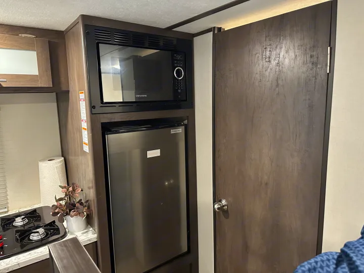 Compact RV rental kitchen with microwave, fridge, and stovetop, next to a dark wooden door.. Coleman 17b 2022
