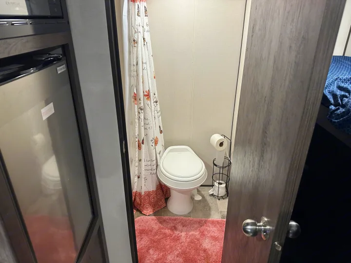 Compact RV rental bathroom featuring a toilet, shower, and soft pink rug, with a built-in kitchen visible nearby.. Coleman 17b 2022