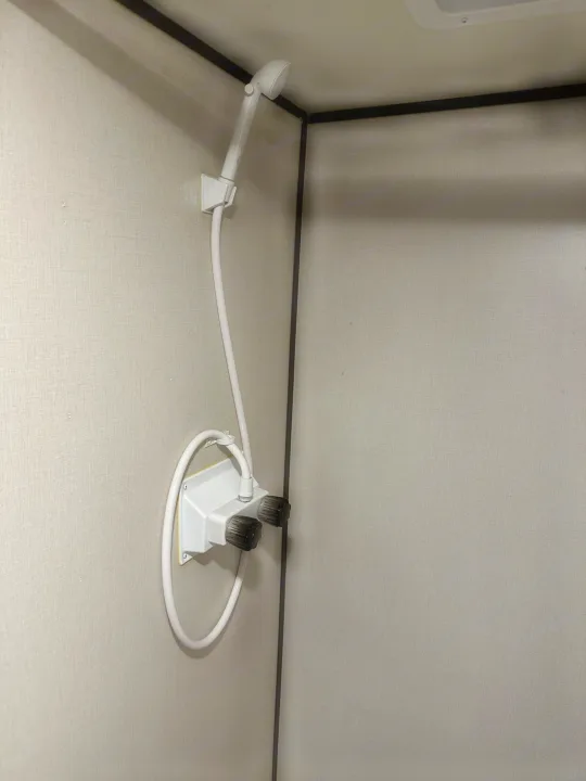RV rental shower inside. A white handheld showerhead is mounted on a light-colored textured wall, with two control knobs below.. Coleman 17b 2022