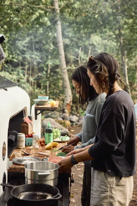 Two women prepare fresh ingredients at an RV rental outdoor kitchen, cooking in a beautiful forest camp setting.. Campworks NS-1 Electrified 2024