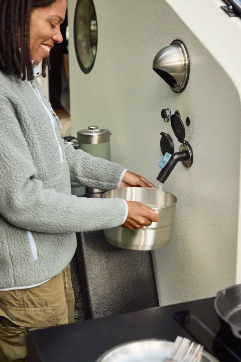 Woman smiles while filling a bowl from a faucet in an RV rental outdoor kitchen, enjoying portable water.. Campworks NS-1 Electrified 2024
