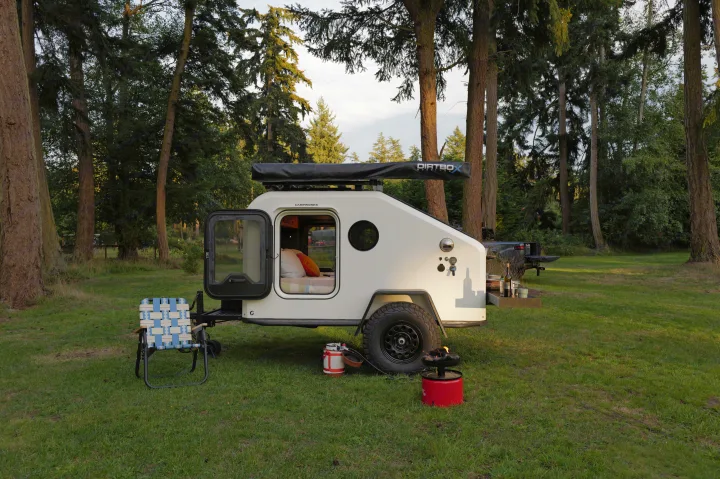 A cozy white teardrop camper, ideal for RV rental adventures, with an open door showing the bed, set up in a lush, wooded campsite.. Campworks NS-1 Electrified 2024