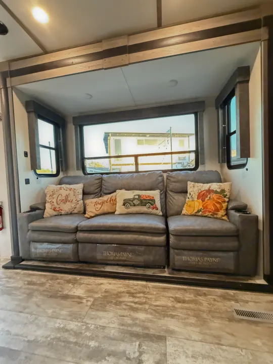 RV rental living area featuring a comfy grey Thomas Payne Collection sofa with fall-themed pillows, large windows, and wood-look floor.. Keystone Carbon 2021
