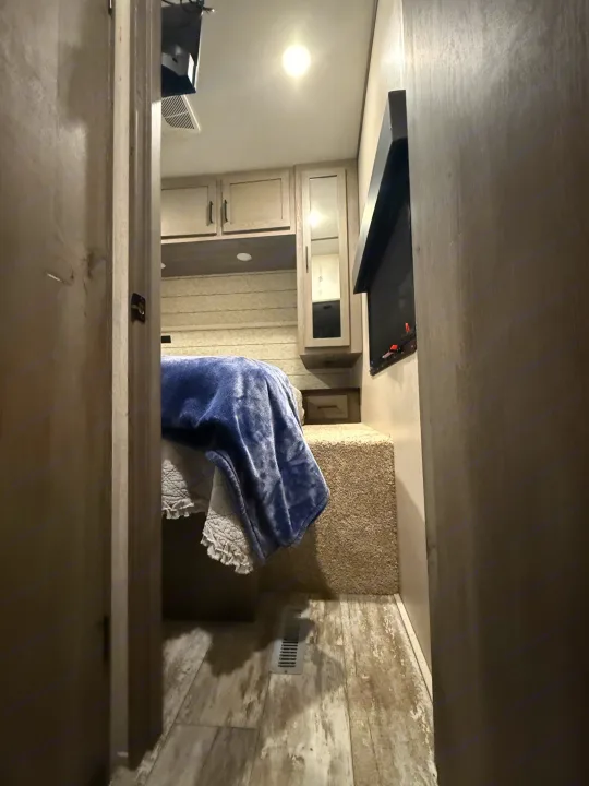 Inside an **RV rental**: a compact bedroom with a blue-blanketed bed, overhead cabinets, and a wall-mounted TV.. Keystone Carbon 2021