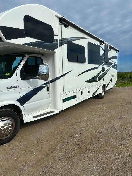 White Class C RV rental, a Jayco Greyhawk, parked on gravel. Its side features black graphics and tinted windows under a cloudy sky.. Jayco Greyhawk 2022