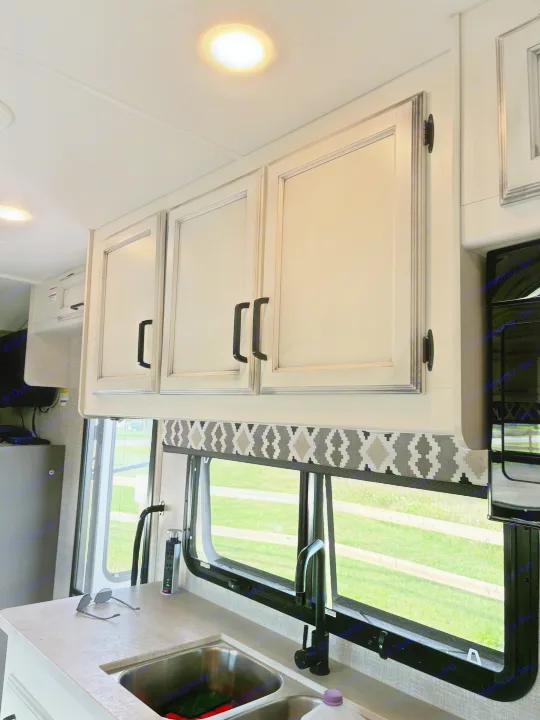 An RV rental kitchen featuring light overhead cabinets with black hardware, a double sink, pop-up outlet, and a window with a patterned valance.. Jayco Greyhawk 2022