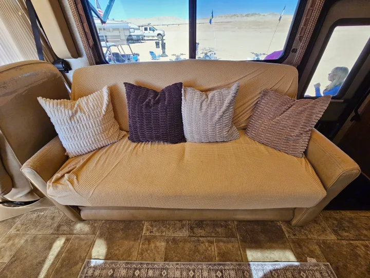 Cozy RV rental interior with couch & pillows. Window view reveals a desert landscape with truck, ATV, and sandy dunes.. Thor Palazzo 2013