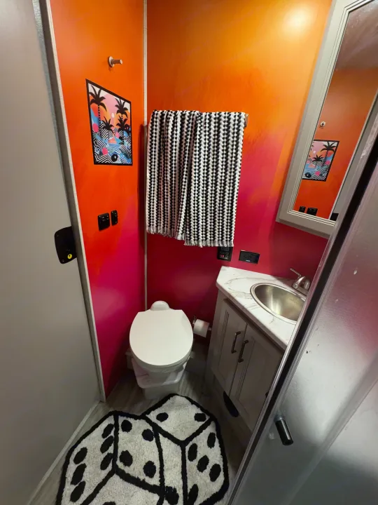 Vibrant RV rental bathroom interior featuring orange-to-pink walls, a toilet, vanity, and quirky dice rug.. Thor Resonate 2024