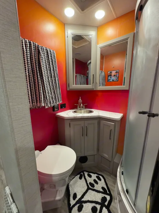 Inside an RV rental bathroom featuring vibrant orange/pink walls, a corner sink, toilet, and shower. A playful dice rug adds character.. Thor Resonate 2024
