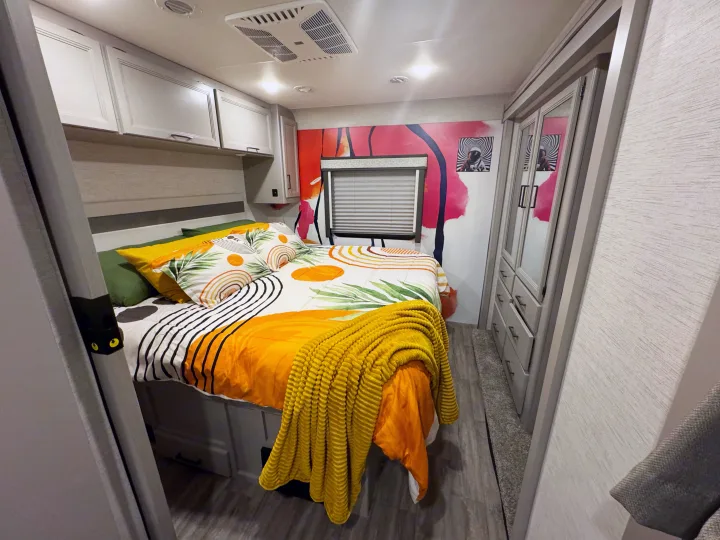 This RV rental bedroom features a vibrant bed, bold wall art, overhead cabinets, and a mirrored wardrobe, creating a stylish space.. Thor Resonate 2024
