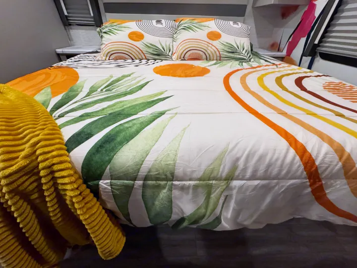 Cozy RV rental bed with modern tropical bedding, featuring green leaves and colorful abstract shapes. A yellow blanket adds warmth.. Thor Resonate 2024