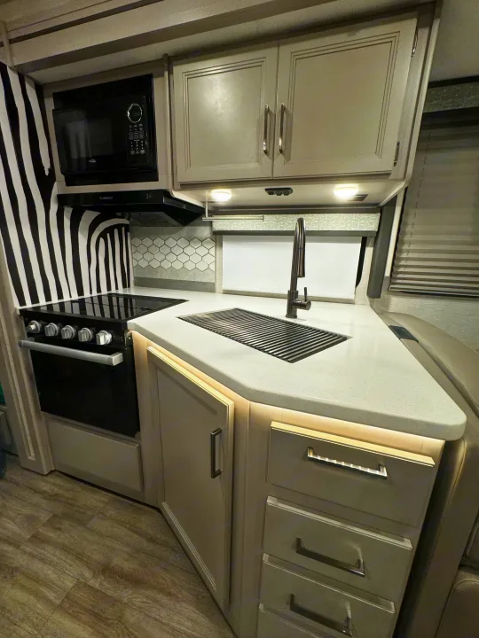 Bright RV rental kitchen: modern gray cabinets, white countertops, microwave, stove, and unique striped wall decor.. Thor Resonate 2024