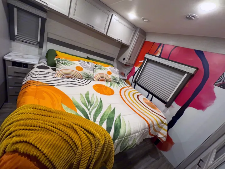 Colorful RV rental bedroom with a bed featuring artistic bedding, a nightstand, overhead cabinets, and abstract wall art.. Thor Resonate 2024