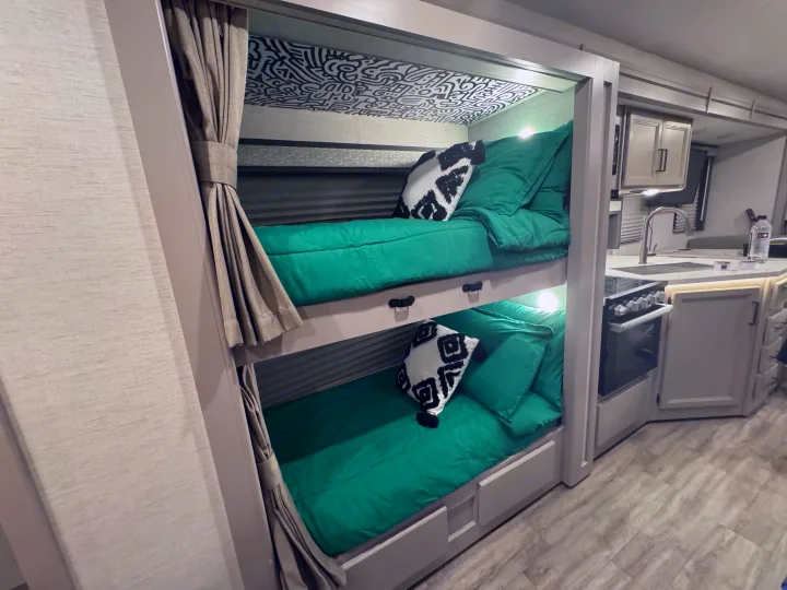 RV rental interior features two cozy bunk beds with bright green bedding, patterned ceilings, and a compact kitchen space.. Thor Resonate 2024